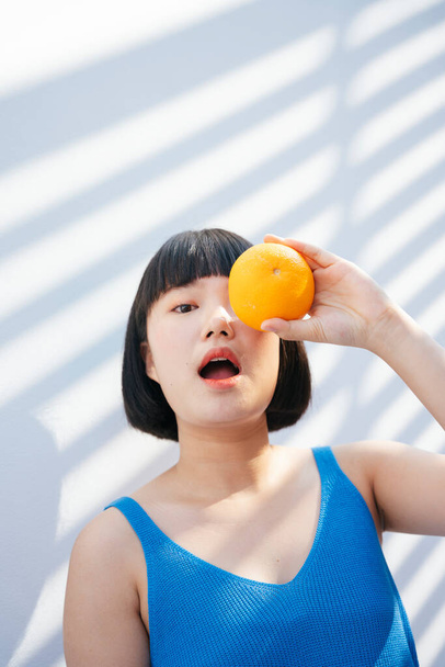 Hermoso corte de pelo bob mujer asiática en camiseta azul tanque sostienen una fruta naranja, abriendo la boca. Listo para comer. Aislado sobre fondo blanco. - Foto, imagen