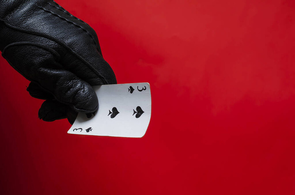Male hand in leather glove holds playing card. The right hand in black glove with triplet of clubs on corral background. Fortune telling or magician concept. Side view. Selective focus. - Photo, image