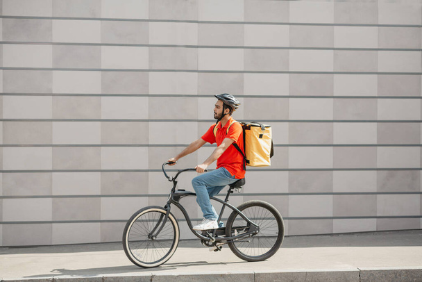 Young caucasian man cyclist in protective helmet with backpack riding bicycle on sidewalk with backpack in city - 写真・画像