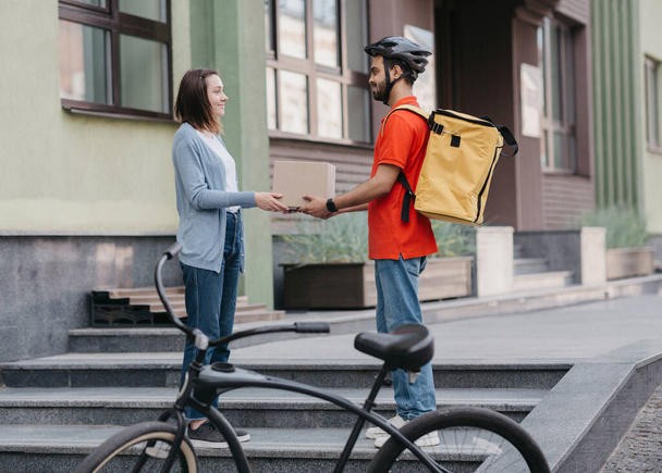 Курьерские услуги и интернет-магазины. Молодой человек с желтым рюкзаком, шлемом и велосипедом дает девушке картонную коробку на улице - Фото, изображение