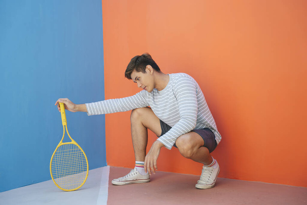 Atrakcyjny młody Azjata w luźnym kostiumie trzymający rakietę tenisową na pomarańczowym tle. Zabawne pozowanie. Gram w tenisa. Siedzenie z gestem rakiety. - Zdjęcie, obraz