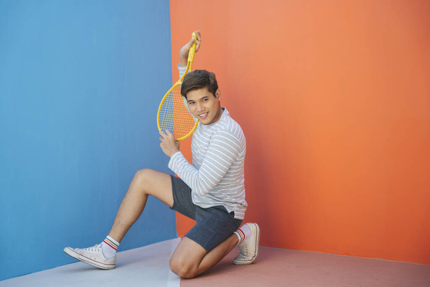 Atrakcyjny młody Azjata w luźnym kostiumie trzymający rakietę tenisową na pomarańczowym tle. Zabawne pozowanie. Gram w tenisa. Gotowy do uderzenia w piłkę.. - Zdjęcie, obraz