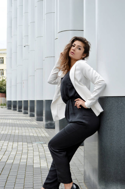 Schöne Geschäftsfrau vor dem Hintergrund des modernen Büros. Mädchen in schwarzen Overalls und weißer Jacke posiert in der Nähe des Business Centers - Foto, Bild