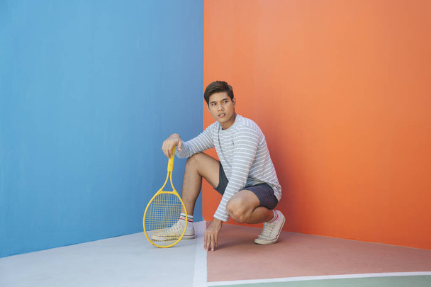 Mavi turuncu arka planda tenis raketi tutan sıradan kostümlü çekici Asyalı bir genç. Komik poz. Tenis oynuyordum. Yerde raket hareketleriyle oturan bir adam.. - Fotoğraf, Görsel