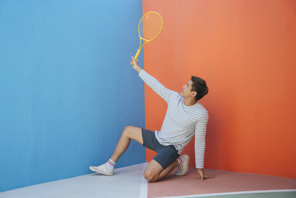 Atraktivní mladý Asiat v neformálním kostýmu drží tenisovou raketu přes modrooranžové pozadí. Vtipné pózování. Hraju tenis. To je moje raketové gesto.. - Fotografie, Obrázek