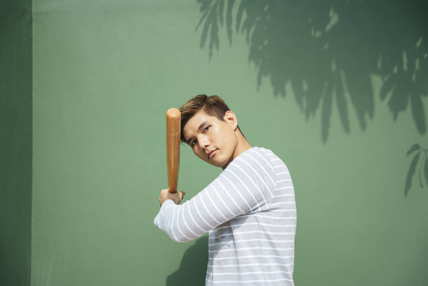 Young handsome asian man hold wooden baseball bat over green background. Baseball player on vacation. - Photo, Image