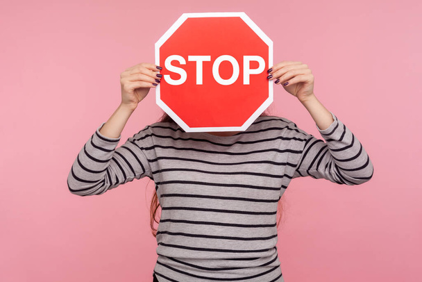 Way prohibited! Portrait of anonymous person in striped sweatshirt covering face with Stop symbol, holding red traffic sign and warning of ban, forbidden access. indoor studio shot, pink background - Photo, Image
