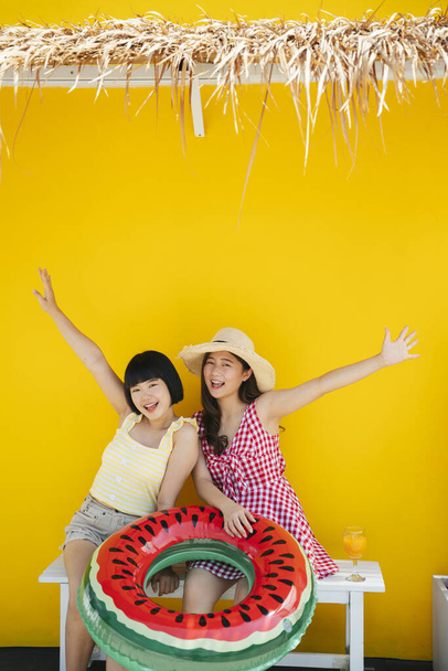Dwie azjatyckie kobiety są bliskimi przyjaciółkami na wakacjach. Podróżując razem po plaży. Na żółtym tle. - Zdjęcie, obraz