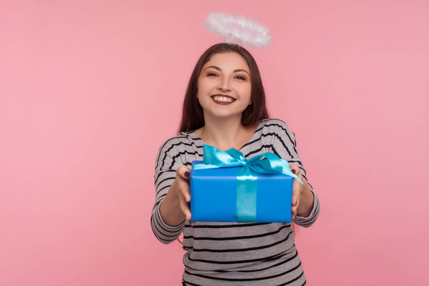 Portrait of angelic girl with saint halo over head giving birthday gift and smiling, sharing present, donating and offering surprise in box, congratulation on anniversary. indoor studio shot isolated - Fotó, kép
