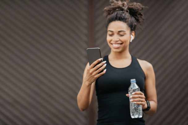 Улыбающаяся африканская американка в спортивной одежде с современными наушниками и фитнес-трекером, смотрит на смартфон и держит бутылку - Фото, изображение