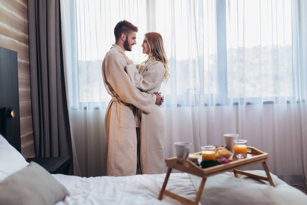 Młoda szczęśliwa para w pokoju hotelowym rano. Tylko żonaty mężczyzna i kobieta stojący przy oknie. - Zdjęcie, obraz