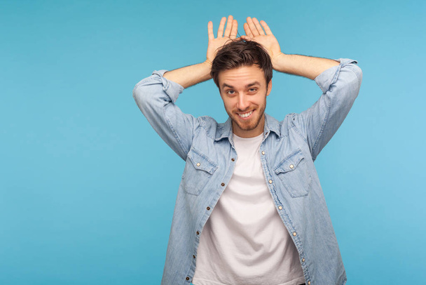 I'm rabbit! Portrait of cheerful carefree handsome man in worker denim shirt showing bunny ears gesture and looking at camera with playful attractive smile. studio shot isolated on blue background - Photo, Image