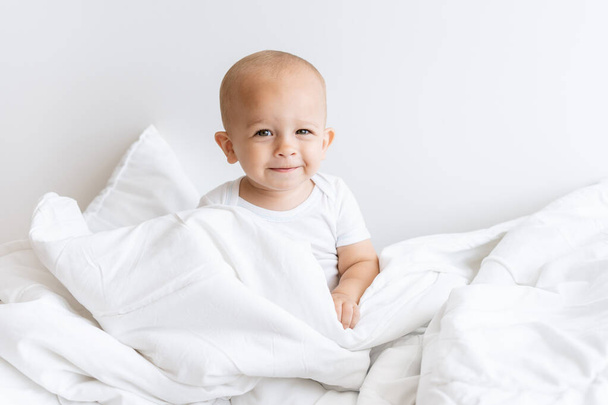 白いベッドの上の白い毛布の中に座っている子供の男の子 - 写真・画像