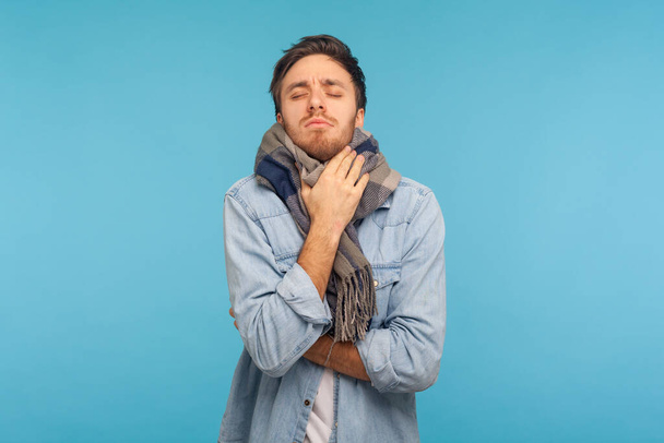 Portrait of unhealthy flu-sick man standing wrapped in warm scarf shivering from cold, feeling unwell suffering fever, seasonal influenza symptoms. indoor studio shot isolated on blue background - Foto, Imagem
