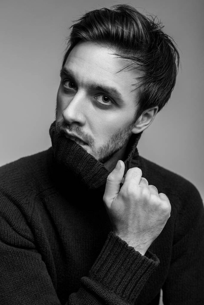 Portrait of sexy handsome man with stylish hairdo wearing warm sweater, biting high collar and looking seductive, flirting at camera, male fashion, vogue concept. black and white, studio shot isolated - Valokuva, kuva