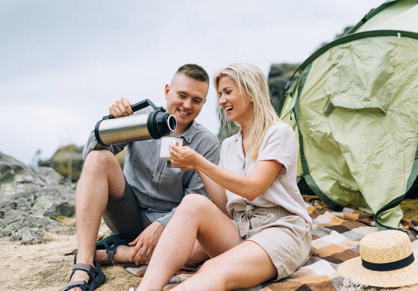 Giovani viaggiatori coppia in abiti casual con tenda. L'uomo versa il tè dal thermos. Turismo locale, fine settimana - Foto, immagini