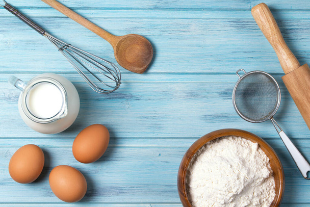 Hozzávalók sütéshez, tejhez, tojáshoz, búzaliszthez és konyhai eszközökhöz kék fa alapon, felülnézetből - Fotó, kép