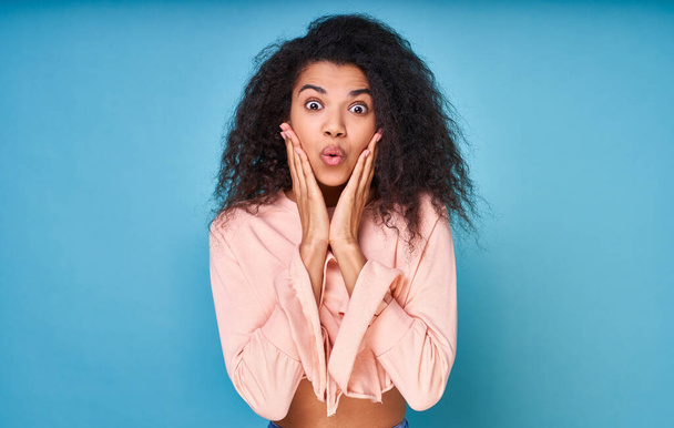 Curly-haired optimistic woman raises her palms in joy, glad to receive an awesome gift from someone, wearing casual beige sweater isolated on blue background. Excited afro woman. Copy space. - Foto, immagini