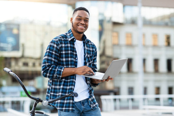 Black man using laptop, standing in downtown with bike - Photo, Image