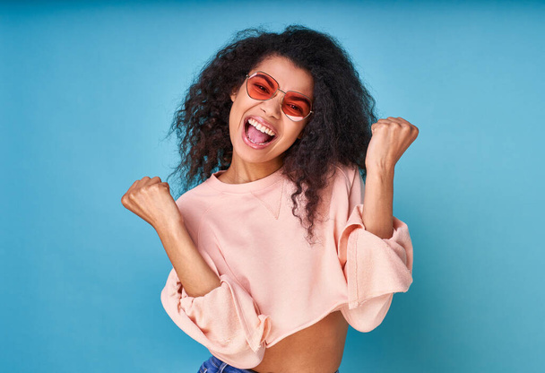 Joyful black lady in beige sweater and red sunglasses, keeps fists clenched, tilts her head and exclaims in triumph, celebrates success, isolated on blue background. - Photo, Image