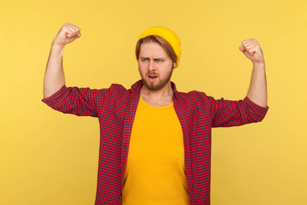 I am strong! Proud hipster bearded guy in beanie hat and checkered shirt showing biceps, feeling energy, confidence and power in hands to win, become successful. indoor studio shot isolated on yellow - Foto, imagen