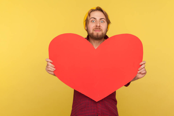 Romantic guy holding big read heart and looking at camera with excitement, demonstrating love affection feelings with huge greeting card on Valentine's Day. studio shot isolated on yellow background - Φωτογραφία, εικόνα