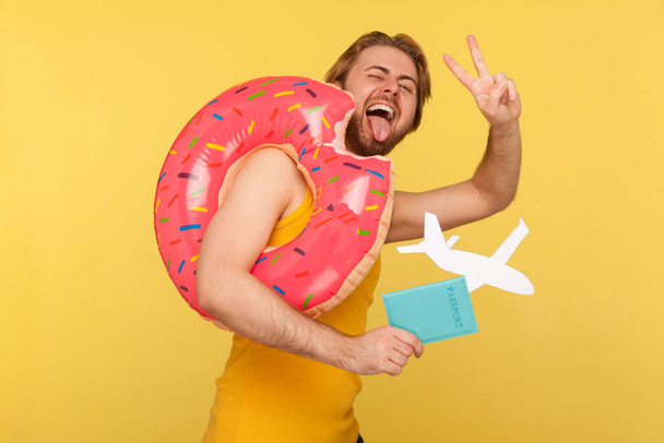 Crazy optimistic tourist guy in undershirt standing with pink donut rubber ring, holding passport document and airplane mockup, showing victory, v sign, gesture of success win. studio shot isolated - Foto, imagen