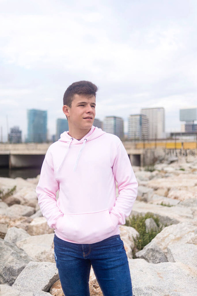 Young teenager male standing against cityscape with hands on pocket on breakwaters while looking away - Photo, Image