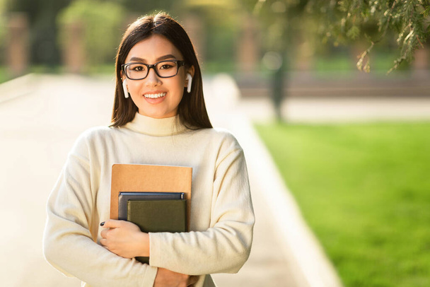 Девушка в очках и наушниках, стоящая в парке с книгами - Фото, изображение