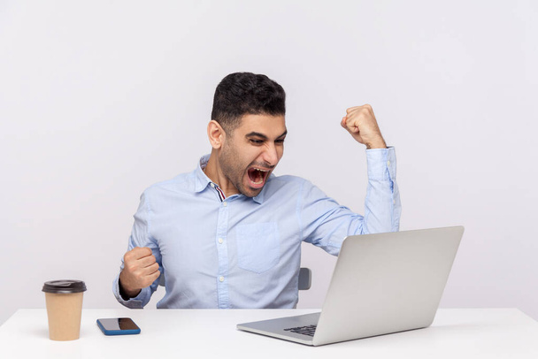 Successful delighted businessman sitting office workplace working on laptop, raising hands shouting for joy and triumphing, celebrating his victory . indoor studio shot isolated on white background - Фото, изображение