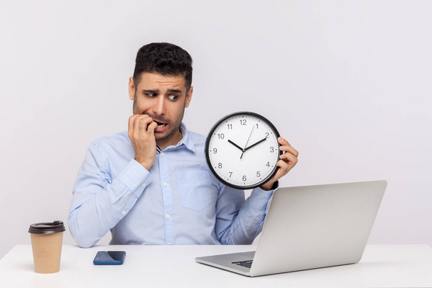 Deadline concept. Nervous anxious man employee sitting in office workplace holding big clock, biting finger nails and looking worried about time limit. indoor studio shot isolated on white background - Фото, изображение
