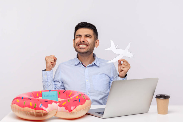 Joyful delighted man holding paper airplane, doing victorious gesture, celebrating travel vacation, weekend trip, sitting in office workplace with rubber ring passport on desk. studio shot isolated - Foto, imagen