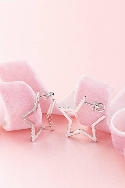 Star shaped silver earring studs decorated with diamonds on pastel pink background with ribbon. Jewelry still life  - Φωτογραφία, εικόνα