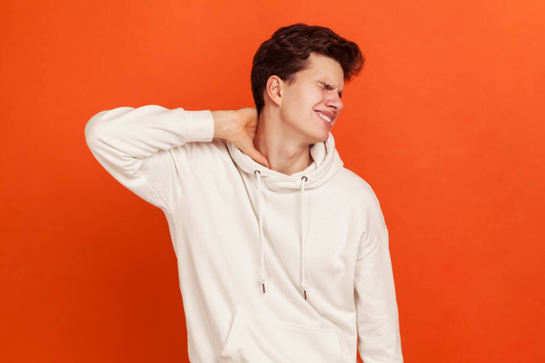 Unhappy worried young man in casual white sweatshirt with hood massaging numb neck, muscle tension, spine injury, osteochondrosis. Indoor studio shot isolated on orange background - Фото, зображення