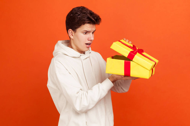 Shocked young man in casual white sweatshirt with hood glancing inside present box, disappointed witt gift, unjustified expectations. Indoor studio shot isolated on orange background - Fotografie, Obrázek