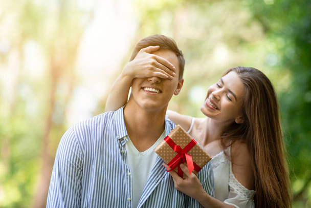 Молодая девушка закрывает глаза своим парням и удивляет его подарком на день рождения в парке - Фото, изображение