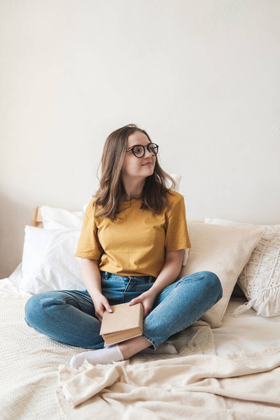 Young beautiful girl student in glasses, an orange T-shirt and blue jeans sits on bed with pillows and reads paper book. Self-education concept at home during quarantine. Home schooling, hobby. - Photo, image