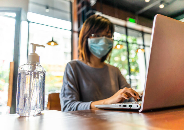 Young asian woman using laptop computer working at home with alcohol gel in quarantine for coronavirus wearing protective mask with social distancing and . Work from home and covid-19 concept. - Фото, зображення