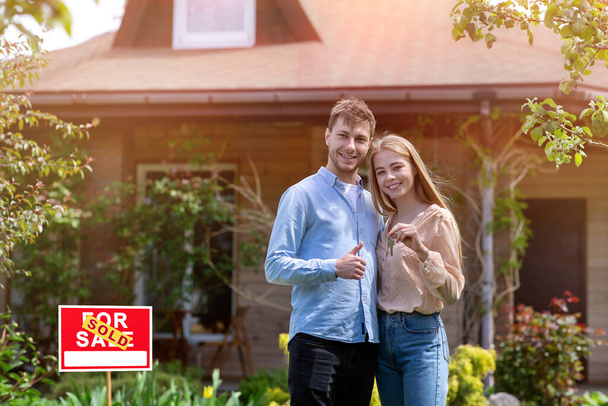 Young lady with house key and her husband showing thumb up in front of their new home, copy space - Photo, Image
