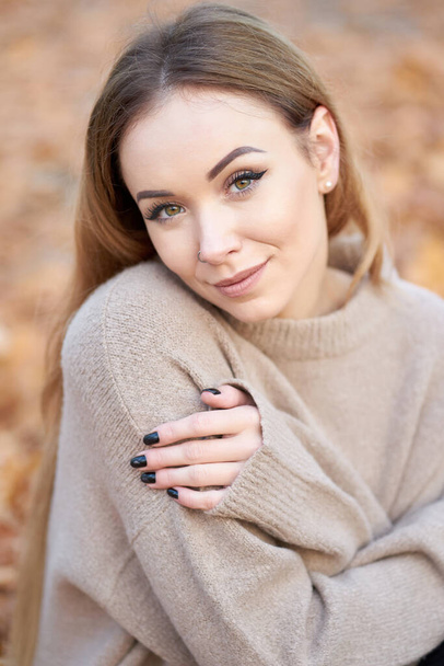 Primo piano ritratto di attraente giovane donna con lunghi capelli biondi indossando accogliente maglione oversize in posa nel parco autunnale.                                - Foto, immagini