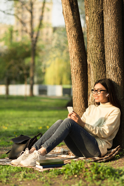 Estudiante usando el teléfono, sentada bajo el árbol en Parkland - Foto, Imagen