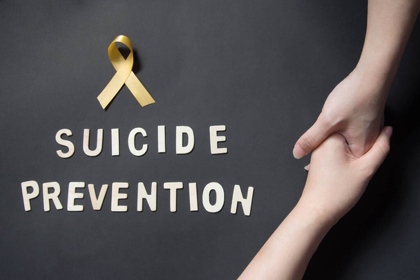 Światowy Dzień Zapobiegania Samobójstwom. Trzymając się za ręce za pomoc i wsparcie depresji kobieta z żółtą wstążką świadomości i SUICIDE PREVENTION drewniane słowo na czarnym tle. Koncepcja zdrowia psychicznego. - Zdjęcie, obraz