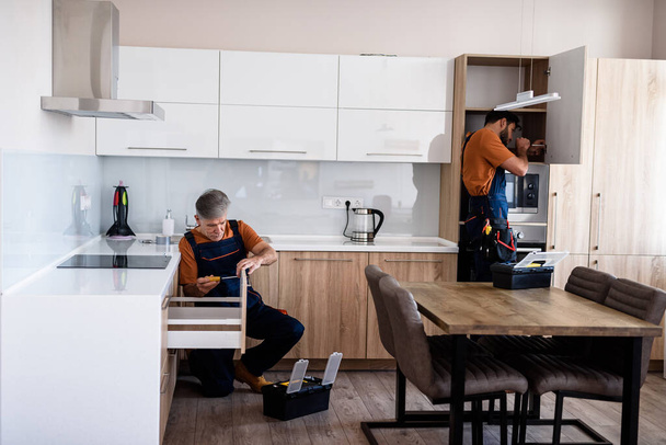 Let us work. Full length shot of two handymen, workers in uniform assembling kitchen cupboard, cabinet using screwdriver indoors. Furniture repair and assembly concept - Foto, immagini
