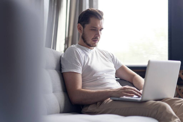 Typing new blog post. Handsome young man using his laptop with smile while sitting on the couch at home. - 写真・画像
