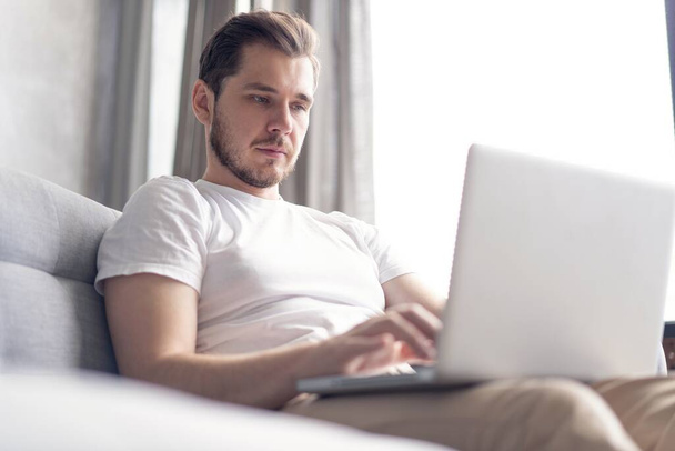 Typing new blog post. Handsome young man using his laptop with smile while sitting on the couch at home. - Foto, imagen