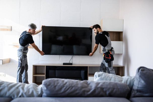 Two professional technicians, workers in uniform installing television on the wall indoors. Construction, maintenance and delivery concept - Photo, Image
