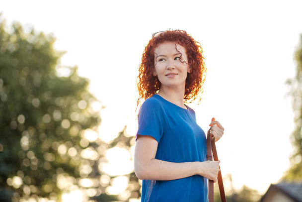 Young woman with waved curly red hair, in blue dress, in park. - Фото, изображение