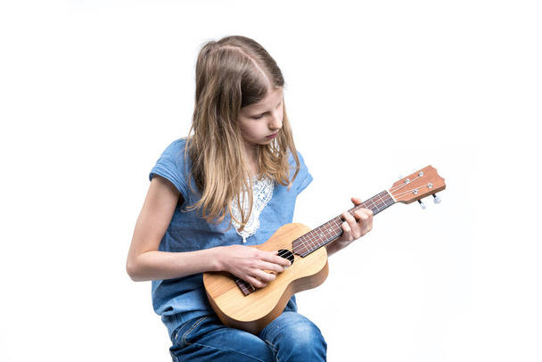 Young, blond girl in blue T-shirt is playing music on ukulele instrument. - Foto, afbeelding