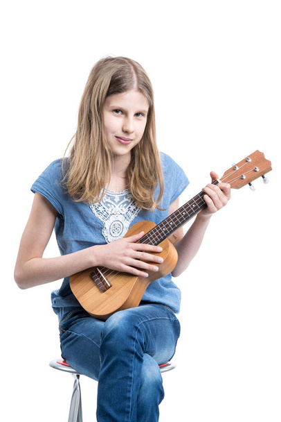 Teenage, blond girl in blue T-shirt is sitting and playing music on ukulele instrument. - Foto, Imagen