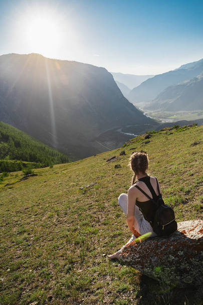 Молода жінка сидить на вершині на тлі красивої долини між горами. Рюкзак пішохідний. Чудовий пейзаж. Наодинці з природою
. - Фото, зображення